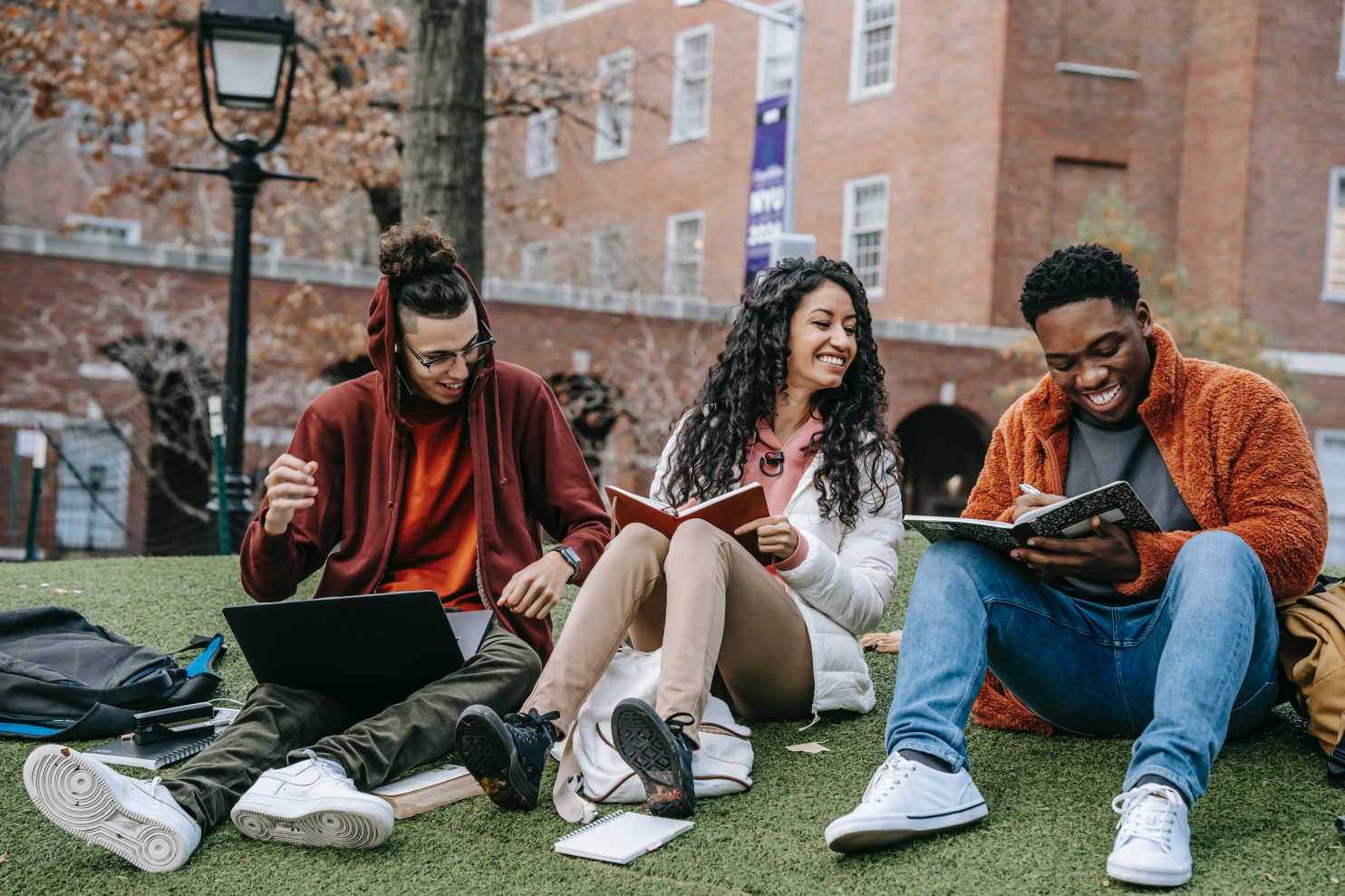 A group of students sitting outside on some grass, looking at books and laughing