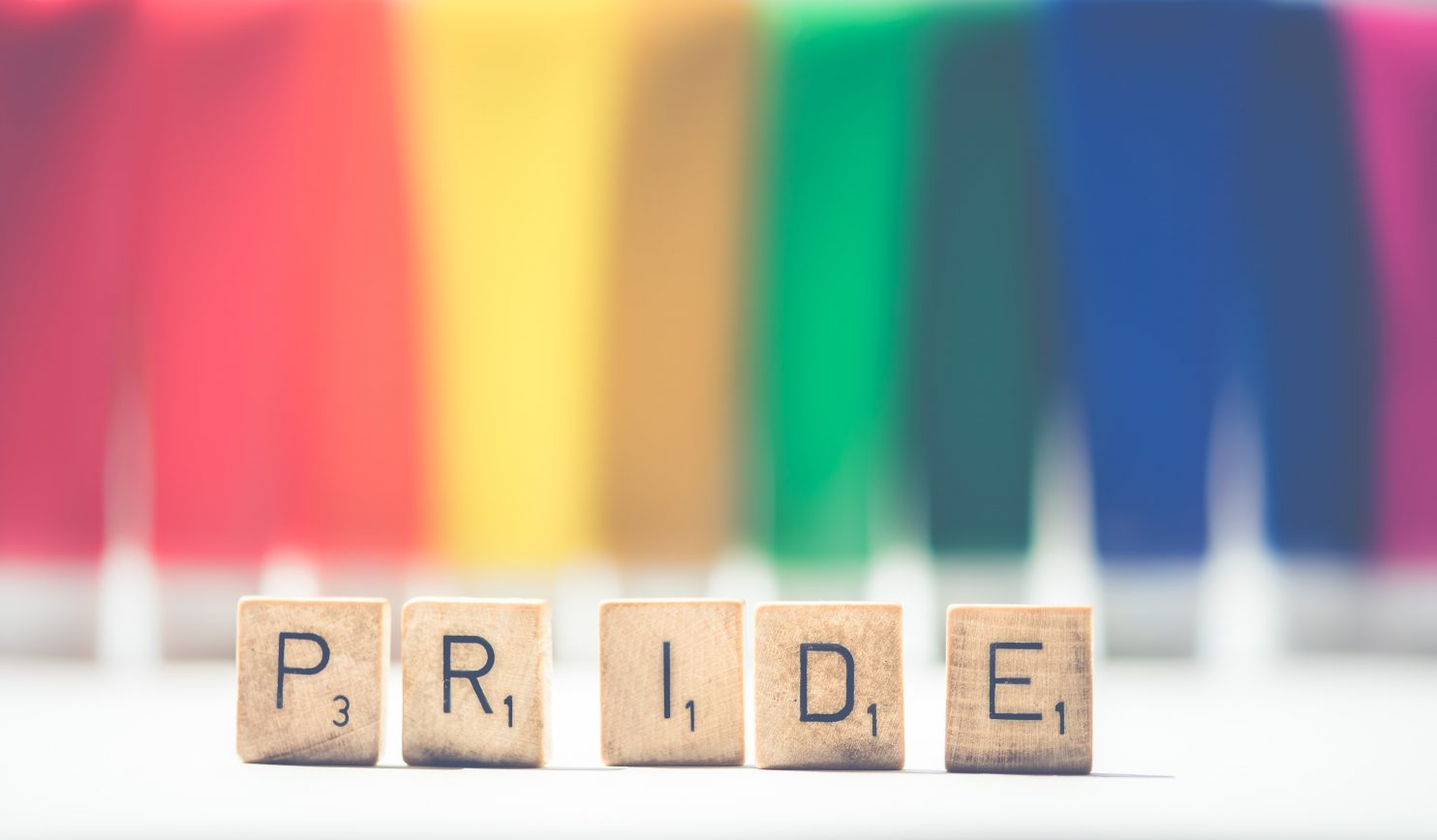 Scrabble tiles that spell out 'Pride' stand in front of a rainbow backdrop